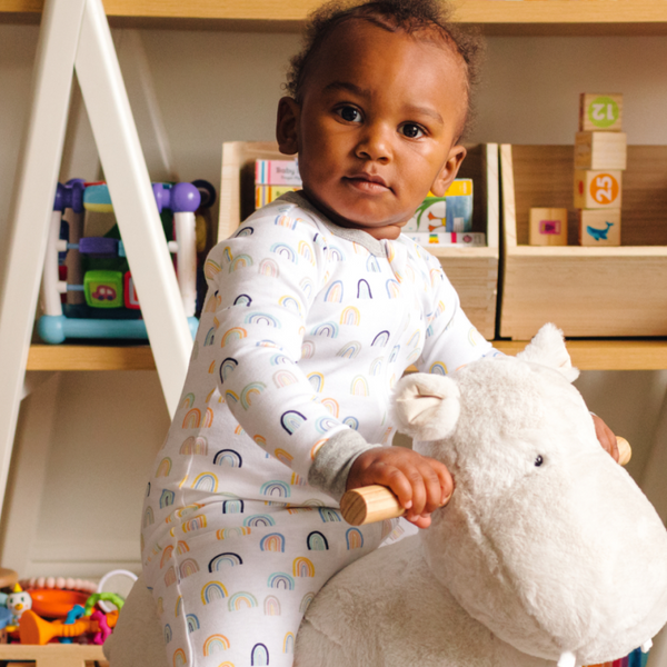 toddler sitting on toy stuffed animal while wearing pajama protective chin zipper guard in sweet rainbows