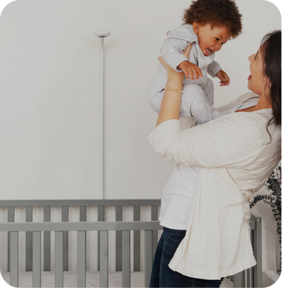 Mom holding happy baby up with view on nanit pro camera and wall mount in background