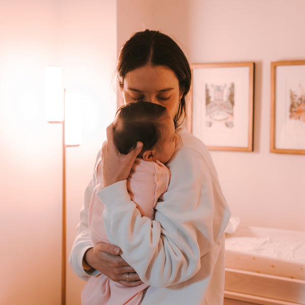 A woman gently holds a baby close in a softly lit room, conveying warmth and tenderness. Framed artwork and a floor lamp are in the background.