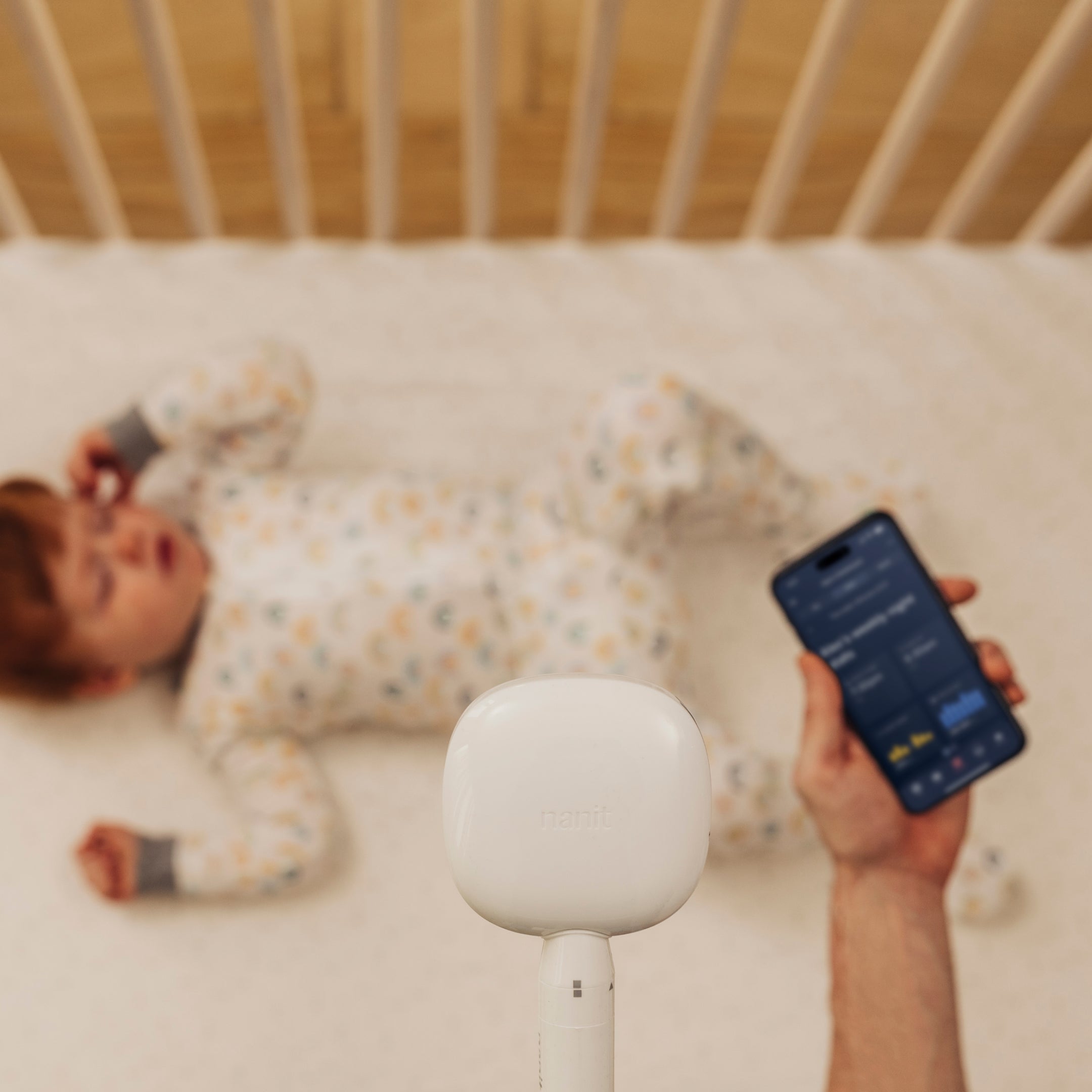 baby in a crib wearing nanit pajamas with nanit pro monitor overhead with person holding mobile phone displaying nanit app