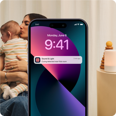 mom holding and kissing baby with sound + light on tableside and phone showing sound + light notification showing "crying detected near kids room"