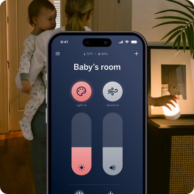 mom holding baby and placing sound + light on side table with phone showing sound + light app with light and sound control and showing temperature and humidity
