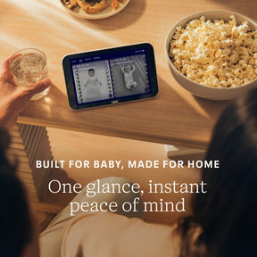 Person holding a glass of water, sitting at a table with a screen displaying a baby monitor, popcorn, and cookies. #complete your setup_camera + screen