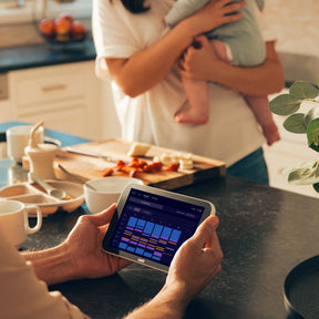 Person holding Nanit Home in a kitchen with a child held by another person.