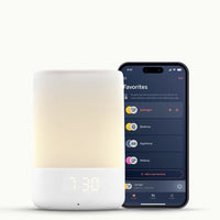 A modern alarm clock with a glowing yellow light and digital time display of 7:30. Next to it is a smartphone showing a sleep app on Nanit app  interface.