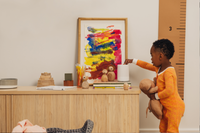 Toddler wearing Nanit Sleep Wear Pajamas in Go Between color going to nanit sound and light machine to change setting with art and home decor in background