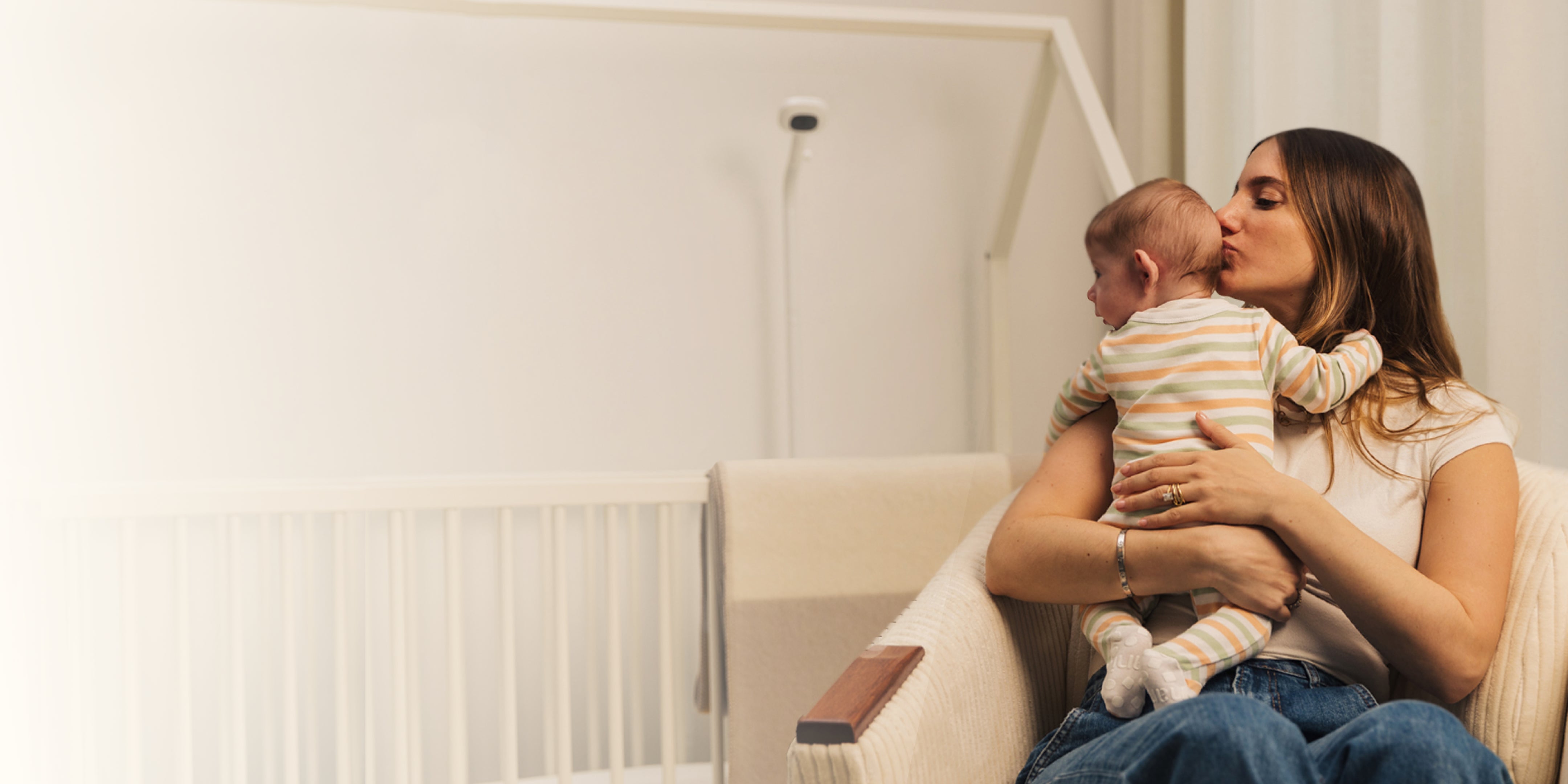 A woman kisses a baby's head while holding them in a cozy chair. The baby wears a striped outfit. The setting feels warm and nurturing.