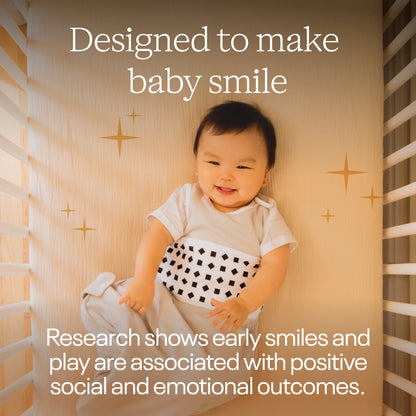 A baby lying in a crib wearing a white outfit with a black-and-white patterned belly band, surrounded by warm text that says 'Designed to make baby smile' and 'Research shows early smiles and play are associated with positive social and emotional outcomes.'.