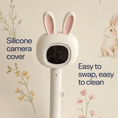 A white bunny-ear shaped silicone camera cover shown on a stand against a light cream wall with floral and rabbit illustrations. Text on the image reads 'Silicone camera cover' and 'Easy to swap, easy to clean'.
