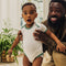 Dad holding baby from behind while baby is standing - baby wearing nanit sleep wear short sleeve bodysuit in spruce green