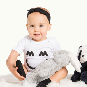 Baby wearing white Miles & Milan Big Smile Bodysuit and wearing a black headband and black sock and holding a rabbit toy