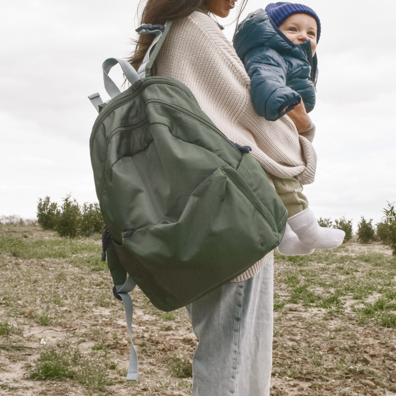 mom holding baby outside while wearing the state lorimer diaper bag in olive